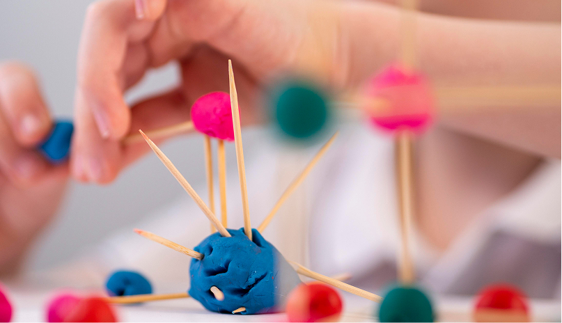 Mano de un niño construyendo una estructura con plastilina y palillos, representando la evaluación psicopedagógica.